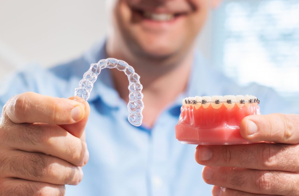 A person holding out an Invisalign clear aligner in one hand and a model of teeth with braces on them in the other hand.