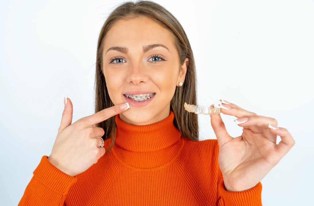 A person pointing at their teeth with braces on them while holding an Invisalign clear aligner in the other hand.