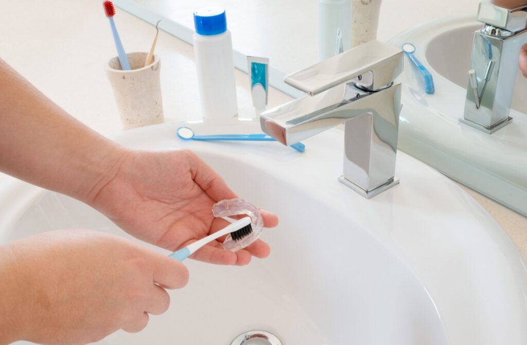A person holding their Invisalign clear aligner under the tap, using a soft bristle tooth brush to clean the aligner.
